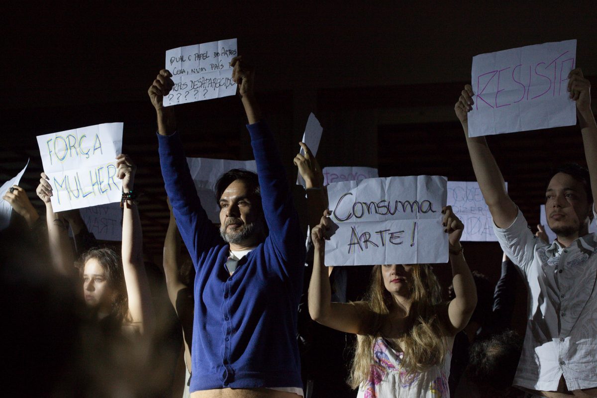 Artistas mostram cartazes com frases de protesto ("Força, Mulher", "Consuma Arte!", "Resista") em encerramento de Festival de Teatro em Bumenau
